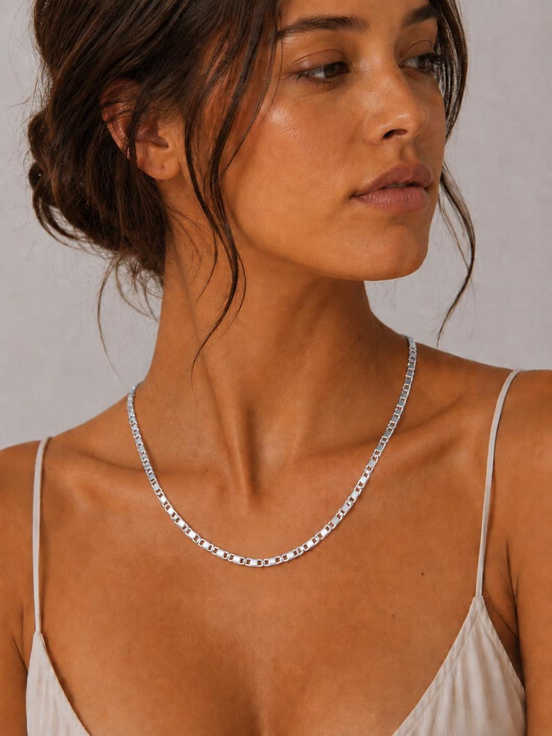 May include: A silver-colored necklace with a flat, rectangular link design. The necklace is worn around the neck and rests on the chest. The model is wearing a white spaghetti strap top.