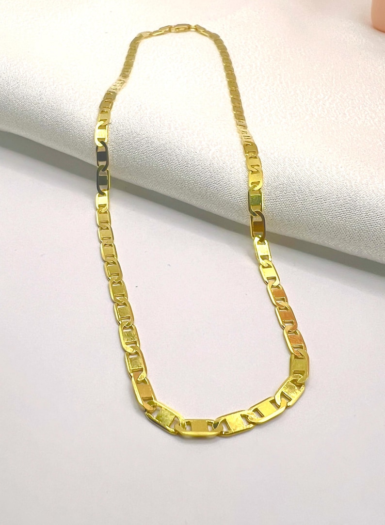May include: A gold-colored chain necklace with a flat, rectangular link design. The necklace is displayed on a white, textured surface. The chain is a bright, shiny gold color.