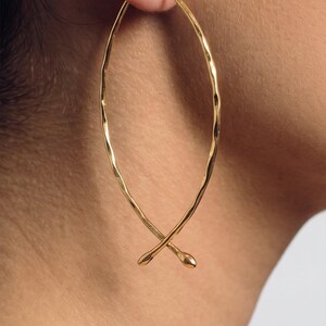 May include: A gold-toned earring featuring an elongated, teardrop-shaped wire frame with a hammered, textured surface. The wire curves upward and crosses at the bottom, ending in two small, rounded, smooth tips that overlap slightly.