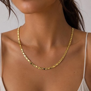 May include: A gold-colored necklace with a flat, rectangular link design. The necklace is worn around the neck and rests on the chest. The chain is made of a shiny, reflective metal.