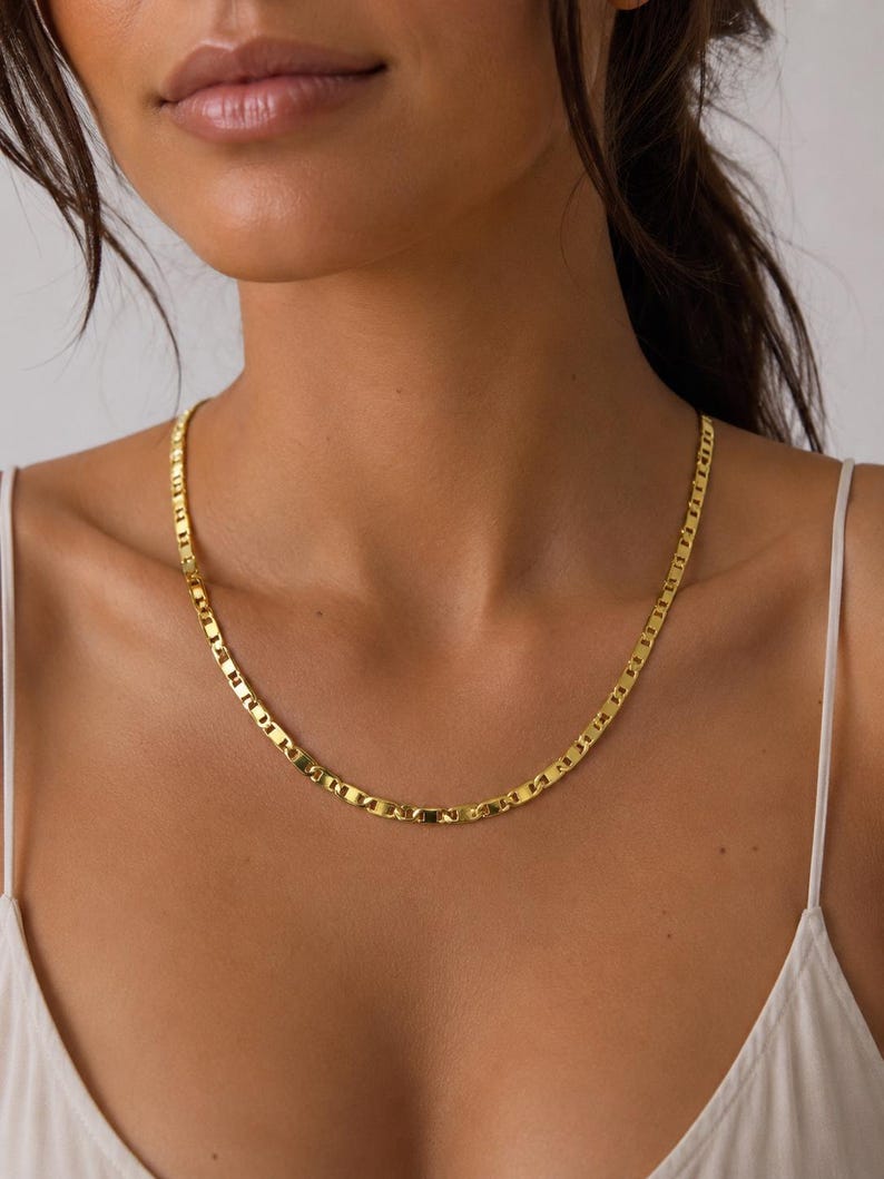 May include: A gold-colored necklace with a flat, rectangular link design. The necklace is worn around the neck and rests on the chest. The chain is made of a shiny, reflective metal.