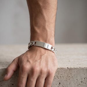 Puede incluir: Una pulsera de identificación de metal plateado con una placa rectangular pulida centrada en una cadena de eslabones gruesos. La pulsera tiene una superficie lisa y reflectante, con una construcción de eslabones que rodea la muñeca.