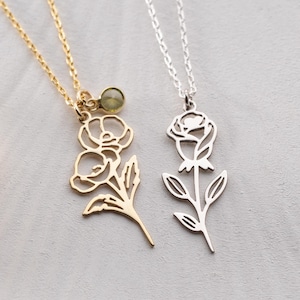 May include: Two delicate necklaces with floral pendants. One is gold-toned with a small green charm, the other is silver-toned. Both feature a chain and a stylized flower design. The necklaces are displayed on a white surface with dried flowers.