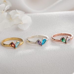 Könnte beinhalten: Drei Ringe mit Edelstein-Akzenten. Die Ringe sind goldfarben, silberfarben und roségoldfarben. Jeder Ring hat zwei tropfenförmige Edelsteine in verschiedenen Farben, darunter Rot, Blau, Lila, Grün und Weiß. Die Ringe werden auf einer weißen, strukturierten Oberfläche präsentiert.