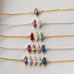 May include: A collection of delicate necklaces with colorful, marquise-cut gemstones. The chains are either gold or silver-toned, and the gemstones include purple, red, white, blue, and pink. The necklaces are displayed on a white surface.
