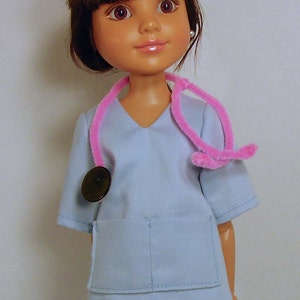 May include: A doll wearing a light blue scrub top with a pink stethoscope and a black button. The doll has brown hair and brown eyes.