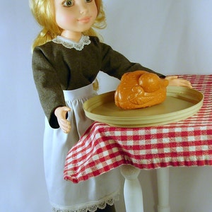 May include: A doll wearing a brown dress with a white apron and lace trim, holding a tray with a brown chicken-shaped toy on a red and white checkered tablecloth.
