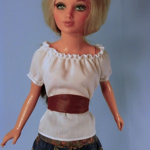 May include: A doll with blonde hair wearing a white peasant top with a brown belt and a blue and green floral skirt.