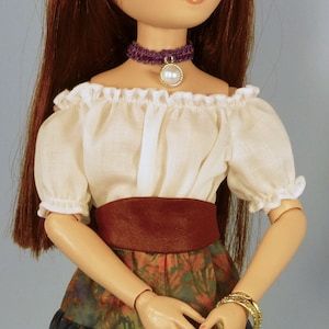 Puede incluir: Una muñeca con un blusa blanca de hombros descubiertos con un cinturón marrón, un collar gargantilla morado con un colgante de perla y una falda de estampado floral. La muñeca tiene el pelo largo y castaño y lleva pulseras doradas en la muñeca.