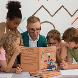 以下が含まれることがあります： 「The Workbook: Helping Anxious Struggling Readers」というタイトルの開かれた本を囲んで、子供たちと大人のグループが集まっています。本はカラフルなイラストで飾られており、他の本が積み重ねられたテーブルの上に置かれています。