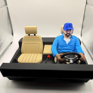 May include: A 3D-printed model of a car interior with a driver. The interior features a tan seat, a black dashboard, and a steering wheel. The driver figure is wearing a blue shirt and a blue cap.
