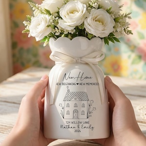 May include: A white ceramic vase with a bouquet of white roses and greenery. The vase has a scalloped rim and a tied ribbon. Text on the vase reads "New Home, New Beginning New Memories", a house illustration, and the address "724 Willow Lane" with names and a year.