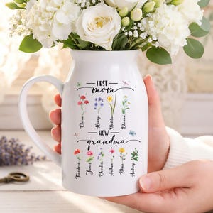 May include: A white ceramic pitcher with a handle, filled with white flowers. The pitcher has the text "First Mom - Now Grandma" with names and floral illustrations. The pitcher is held by a person.