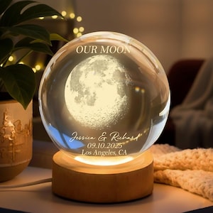 May include: A spherical glass ornament featuring a detailed moon design, lit from within. It rests on a wooden base that provides a warm light. The text "OUR MOON" is at the top, with names and a date below.