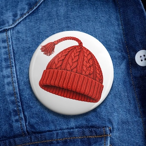 May include: A round, white button featuring a detailed illustration of a red knit beanie with a braided design and a tassel. The beanie is set against a denim background.