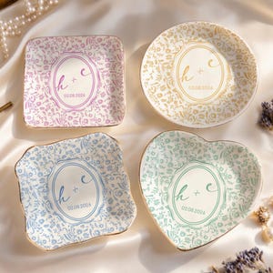 May include: Four decorative ceramic dishes in various shapes: square, round, and two with scalloped edges, one heart-shaped. Each has a floral pattern and a central design with initials and a date. Gold trim edges.