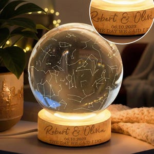 May include: A glowing glass sphere displaying a constellation pattern, resting on a wooden base. The base is engraved with the names "Robert & Olivia", the date "06.10.2023", and the words "THE DAY WE SAID I DO". The sphere emits a warm light.