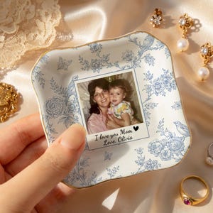 May include: A square, white ceramic trinket dish with a blue floral pattern and gold trim. The dish holds a photo with the text "I love you Mom Love, Olivia". Decorative jewelry is in the background.