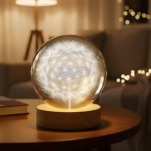 May include: A decorative lamp featuring a clear glass sphere with a detailed dandelion design, illuminated with a soft, warm light. The sphere sits on a round, wooden base, creating a cozy and inviting ambiance. The lamp is on a wooden table.