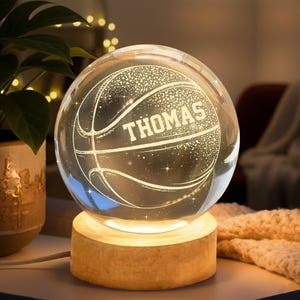 May include: A clear glass basketball-shaped night light with the name "THOMAS" etched on the front. The basketball design is illuminated with white light and sits on a wooden base. The light is plugged in.