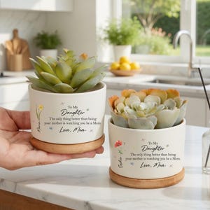 May include: Two small white ceramic planters with succulents, each on a wooden base. The planters have floral designs and text that reads "To My Daughter" and "Love, Mom." One is held by a hand.