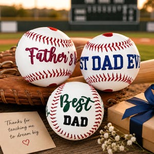 May include: Three white baseballs with red stitching and text that reads "Father's", "Best Dad Ever", and "Best Dad". A baseball glove, a baseball bat, a gift box with a blue ribbon, and a card that says "Thanks for teaching me to dream big" are also in the image.