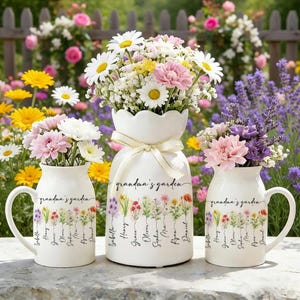 May include: Three white ceramic vases filled with colorful flowers. The vases are decorated with floral designs and the text "grandma's garden". The largest vase has a scalloped top and a ribbon. The other two vases have handles.