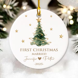May include: White ceramic Christmas ornament with a gold string. The ornament features a green watercolor Christmas tree with a gold star on top, surrounded by gold stars. Text reads "FIRST CHRISTMAS MARRIED" and names "Jennifer ❤ Peter" with the year "-2025-".