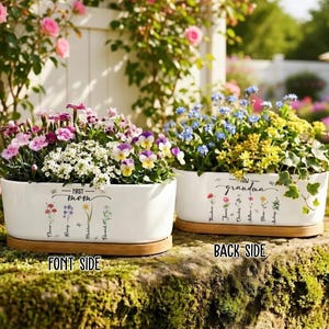 May include: Two white ceramic planters filled with colorful flowers. One planter says "First Mom" and the other says "Grandma". Each planter sits on a wooden base. The planters are outdoors with a background of flowers.