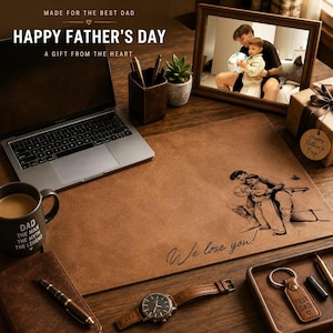 May include: A desk setup with a laptop, coffee mug, and watch. A framed photo and a gift box are present. A leather desk mat features an engraved image and the words "We love you!" The text "Happy Father's Day" is visible.