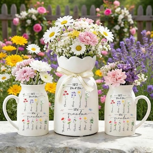 May include: Three white ceramic vases filled with colorful flowers. The vases are decorated with the words "First Mom, Now Grandma" and floral illustrations. The largest vase has a scalloped top and a ribbon. The other two vases have handles.