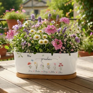 May include: A white ceramic planter filled with a variety of colorful flowers, including daisies, lavender, and pink blooms. The planter has the word "grandma" and names of loved ones. It sits on a wooden base, perfect for a garden or patio.