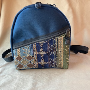May include: A small navy blue backpack with a zippered pocket on the front. The pocket is made of a patterned fabric with blue, green, and brown colors.