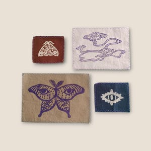 May include: Four fabric patches with purple designs. One patch features a moth, another has cloud-like shapes, a third displays a butterfly with eye-like patterns, and the last shows an eye symbol.