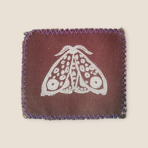 May include: A square, burgundy fabric patch with a white moth design. The moth has a detailed wing pattern. The edges are stitched with purple thread. The patch has a rustic, handmade appearance.