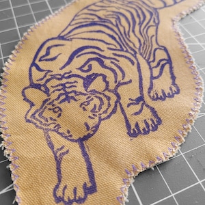 Può includere: Una toppa in tessuto beige con un disegno di tigre blu. La tigre è raffigurata in posizione accovacciata, con strisce e lineamenti del viso dettagliati. La toppa ha un bordo cucito viola, che le conferisce un aspetto fatto a mano.