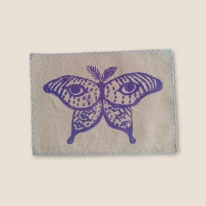 May include: A rectangular patch featuring a purple butterfly design. The butterfly has detailed wings with eye-like patterns, printed on a beige fabric with a blue stitched border. The patch is likely for sewing or ironing onto clothing.