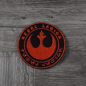 May include: A black and red embroidered patch featuring the Rebel Alliance symbol and the text "Rebel Legion" in a circle.