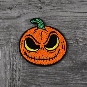May include: Embroidered orange pumpkin patch with a green stem and a menacing face. The patch features black eyes, a stitched mouth, and yellow highlights, perfect for Halloween or spooky-themed projects.