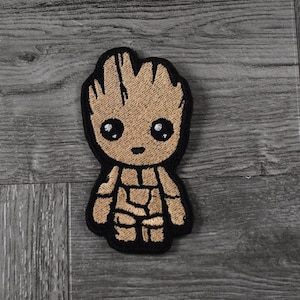 May include: Embroidered patch of a brown cartoon character with a large head and small body. The character has a large smile and large eyes. The character is standing with its arms at its sides. The patch is outlined in black.
