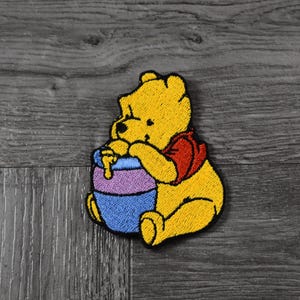 Puede incluir: Un personaje bordado de Winnie the Pooh sosteniendo un tarro de miel morado y azul.