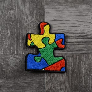 May include: A colorful puzzle piece patch with red, yellow, blue, and green sections. The patch is made of felt and is embroidered with a puzzle piece design.