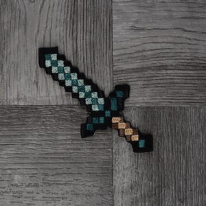 May include: A green and black pixelated sword with a brown wooden handle. The sword is embroidered on a grey fabric background.