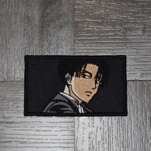 May include: Black and white embroidered patch featuring a close-up of an anime character with brown hair and eyes. The character is wearing a dark suit and has a serious expression.