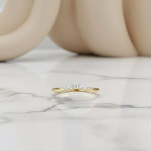 May include: A curved gold ring with five small, clear gemstones set across the top. The ring is set against a white marble surface with a blurred, neutral-toned background.