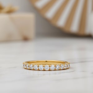 Könnte beinhalten: Ein goldener Bandring mit zahlreichen kleinen, klaren Edelsteinen. Der Ring ist in der Mitte des Bildes platziert, mit einem unscharfen Hintergrund in neutralen Tönen. Das Design des Rings ist schlicht und elegant, passend für verschiedene Anlässe.