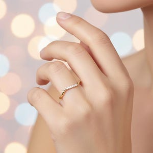 May include: A gold-colored ring with a curved design, set with small, clear gemstones. The ring is worn on a finger, showcasing its delicate and elegant style. The ring's design is a wave-like shape, with the gemstones set along the curve.