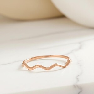 May include: A rose gold ring with a wavy design. The ring is thin and delicate, with a smooth, polished finish. The ring is set against a white marble surface.