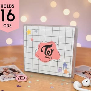 Puede incluir: Una caja blanca para guardar CD con un patrón de cuadrícula y la palabra "TWICE" en un diseño rosa. La caja tiene capacidad para 16 CD y tiene pegatinas decorativas de estrellas y flores. También se ve una tarjeta de fotos y auriculares.