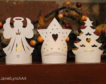 Pode incluir: Três porta-velas de papel branco com desenhos recortados de um anjo, uma estrela e uma árvore de Natal. Cada porta-velas tem um pequeno coração recortado e uma vela de chá dentro.